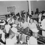 Line at lunch in the cafeteria. The sign on the back wall reads: "Food is exceptionally important in the war effort and must not be wasted. All men are directed to take all the food they want and eat all the take. There is not excuse for food waste. Men wasting food will be subject to disciplinary action."
